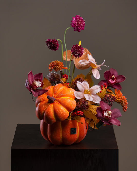 Whimsical Pumpkin Flower Arrangement featuring luxury deep plum orchids, scabiosa, and autumn blooms in an orange pumpkin ceramic vase on a pedestal.