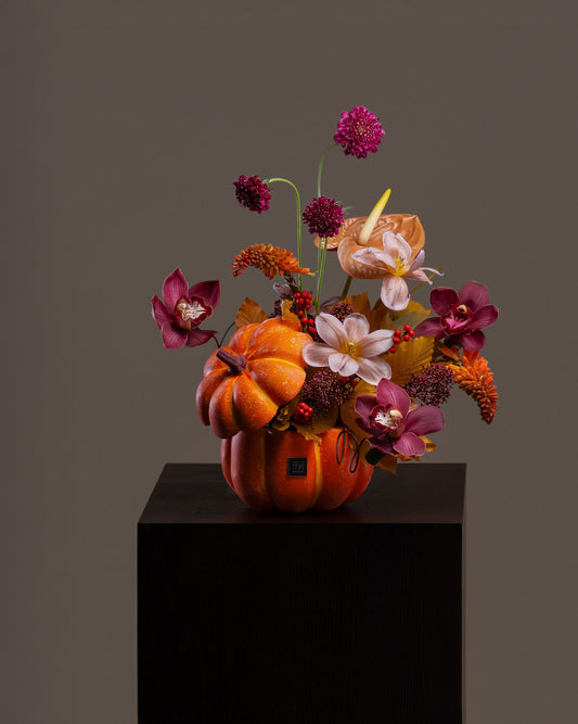 Whimsical Pumpkin Flower Arrangement featuring luxury deep plum orchids, scabiosa, and autumn blooms in an orange pumpkin ceramic vase on a pedestal.